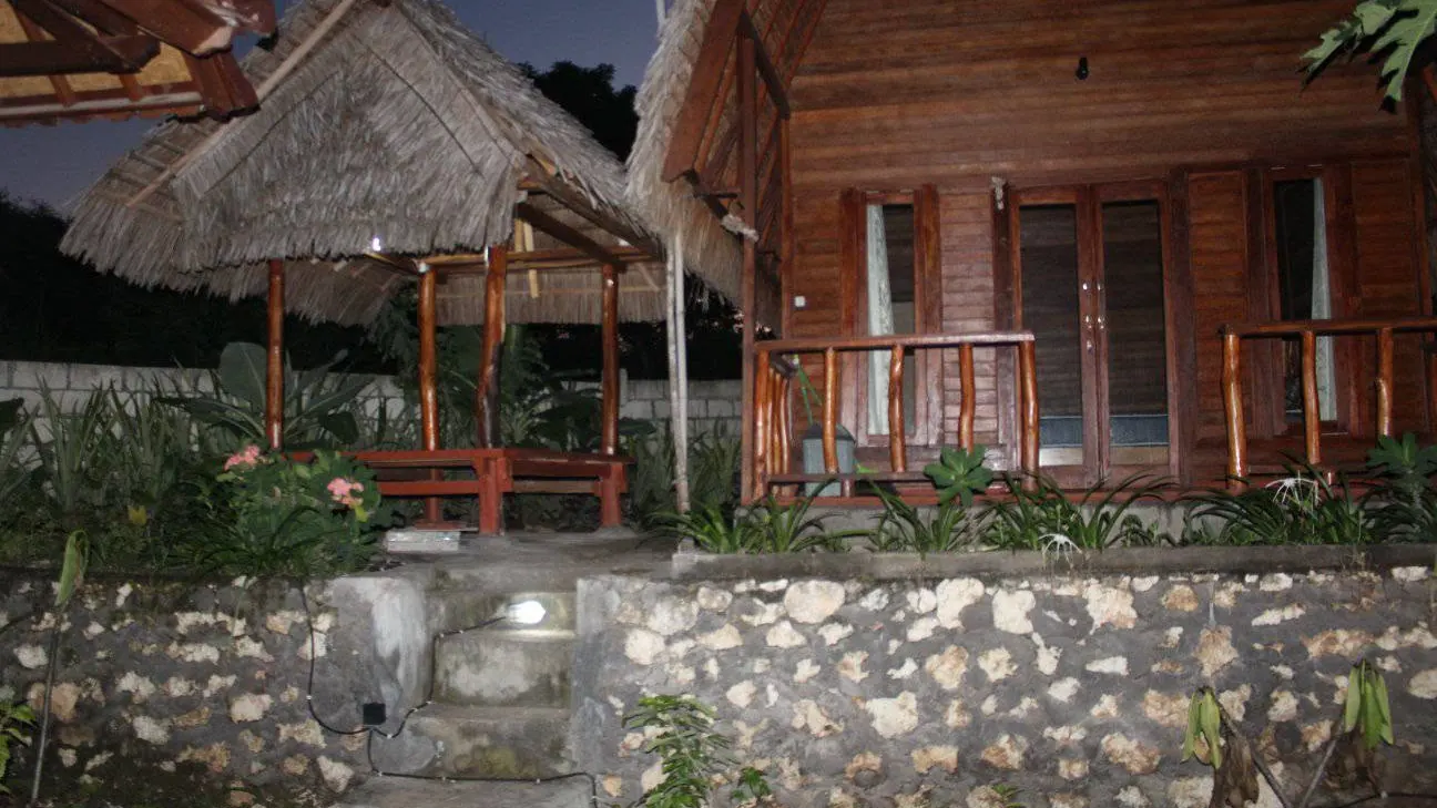 A thatched shelter and a timber cabin behind a low stone wall in the evening